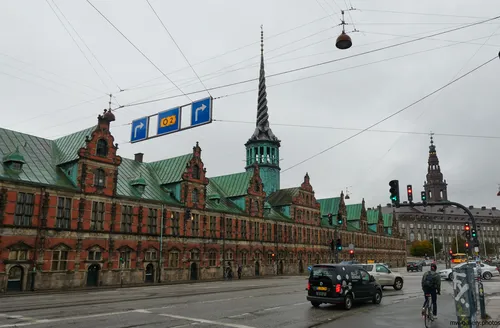The image shows the Copenhagen old stock exchange, known as Børsen, captured during a cloudy morning in autumn. The photograph is an urban architecture shot that highlights the historic building’s distinctive green copper roof and iconic spire, situated in the heart of the city. In the background, the tower of Christiansborg Palace, another recognizable landmark in Copenhagen, rises above the urban streetscape. The busy intersection, traffic lights, and people on bikes further emphasize the city’s vibrant urban atmosphere. This architectural scene showcases Copenhagen’s rich history and city life in the autumn season.